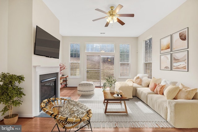 living area featuring baseboards, visible vents, a ceiling fan, a glass covered fireplace, and wood finished floors