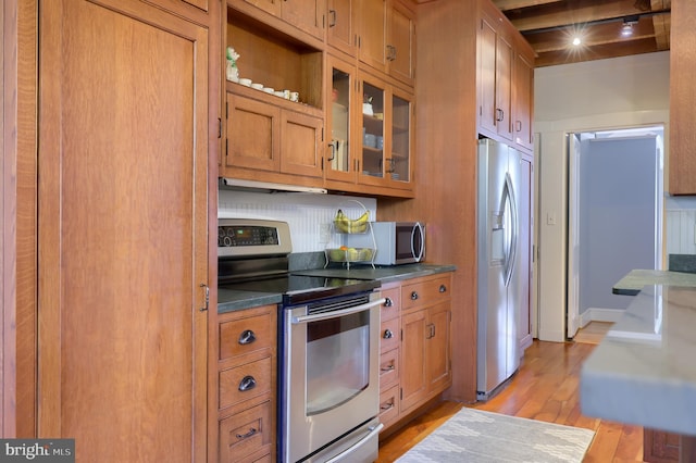 kitchen featuring dark countertops, glass insert cabinets, appliances with stainless steel finishes, light wood-style floors, and brown cabinetry