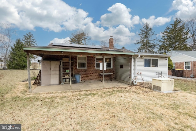 rear view of property featuring a patio, solar panels, brick siding, a lawn, and a chimney