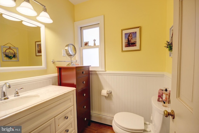 half bath featuring a baseboard radiator, toilet, wood finished floors, vanity, and wainscoting