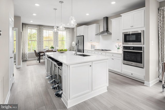 kitchen featuring wall chimney range hood, a center island with sink, light countertops, stainless steel appliances, and a sink