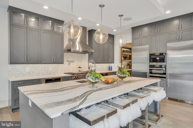 kitchen with stainless steel appliances, wall chimney range hood, light stone counters, and gray cabinetry