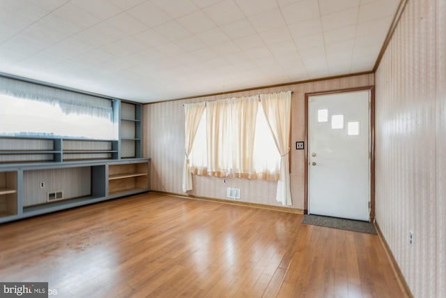 unfurnished living room with wood finished floors, visible vents, baseboards, wallpapered walls, and crown molding