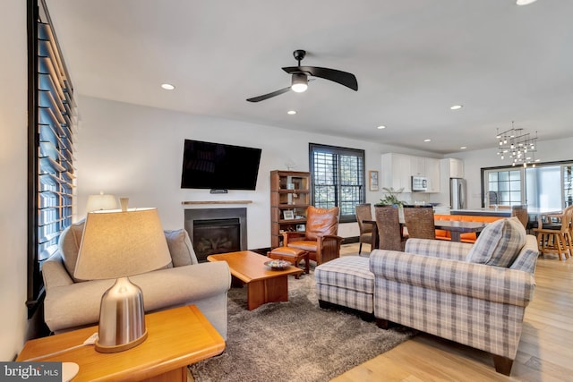 living area with ceiling fan with notable chandelier, light wood finished floors, a glass covered fireplace, and recessed lighting