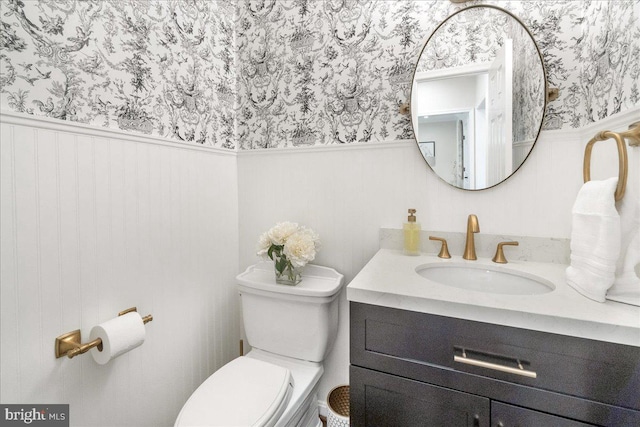 half bathroom with toilet, wallpapered walls, vanity, and wainscoting