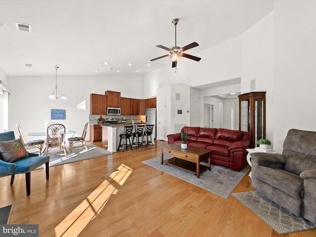 living area with visible vents, a towering ceiling, light wood finished floors, and ceiling fan with notable chandelier