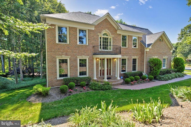 georgian-style home with a front yard, brick siding, a balcony, and roof with shingles