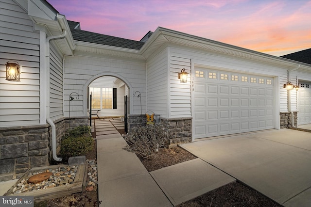 exterior entry at dusk featuring an attached garage, stone siding, concrete driveway, and roof with shingles