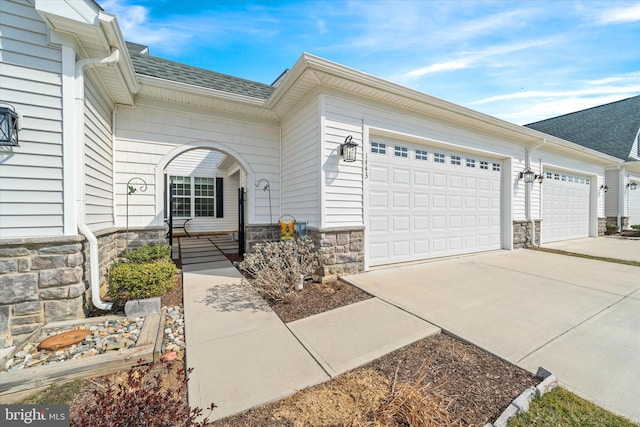 entrance to property featuring stone siding, roof with shingles, an attached garage, and concrete driveway