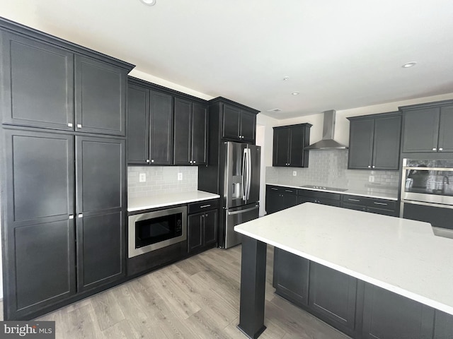 kitchen featuring light countertops, appliances with stainless steel finishes, light wood-style floors, dark cabinets, and wall chimney exhaust hood