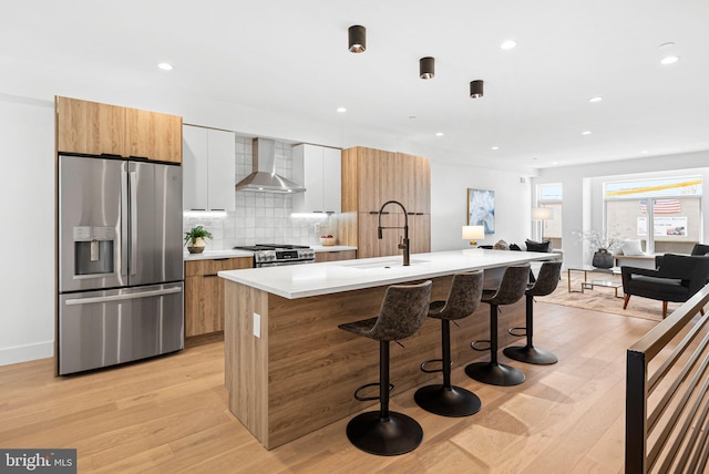 kitchen with an island with sink, a sink, appliances with stainless steel finishes, wall chimney exhaust hood, and modern cabinets