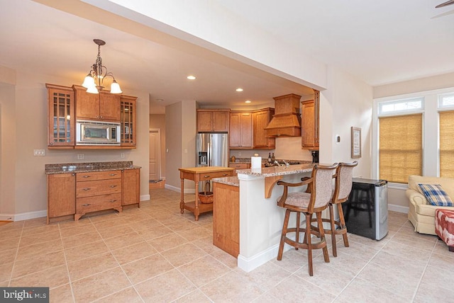 kitchen with stainless steel appliances, brown cabinetry, premium range hood, a peninsula, and a kitchen bar