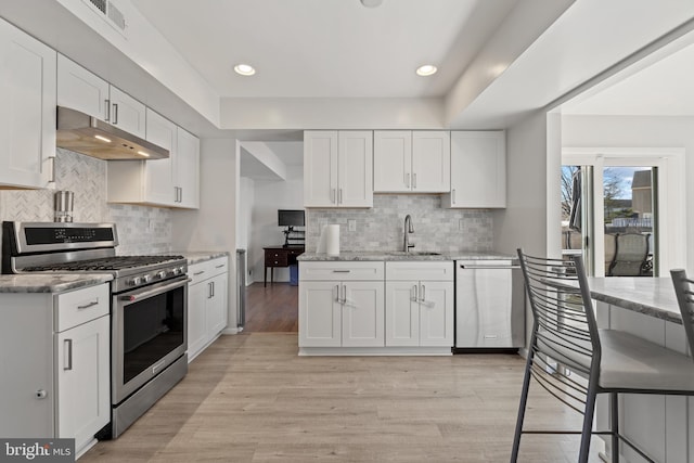 kitchen with under cabinet range hood, a sink, white cabinets, appliances with stainless steel finishes, and light wood finished floors