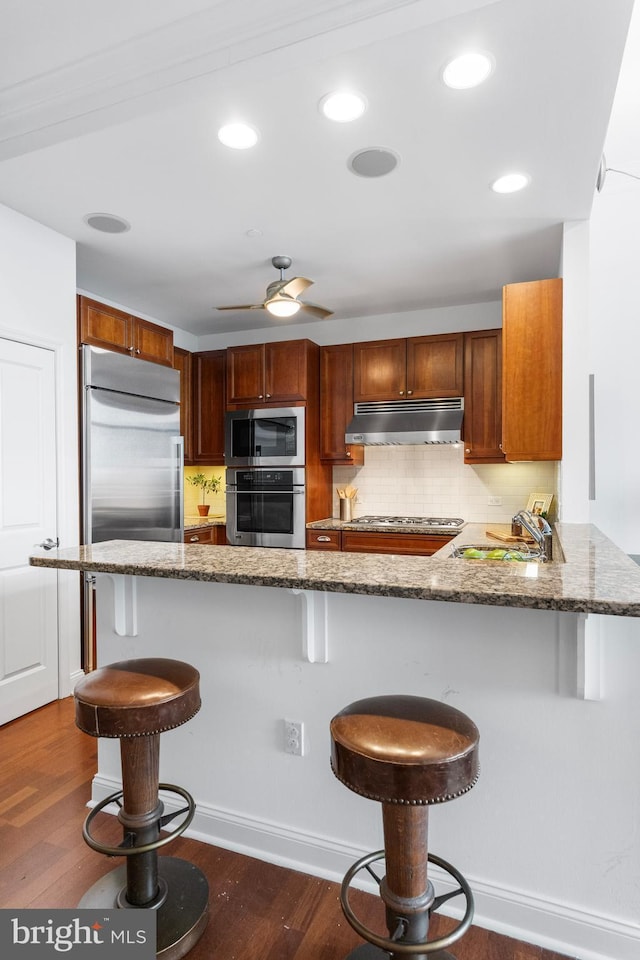 kitchen with built in appliances, stone countertops, a kitchen breakfast bar, and under cabinet range hood