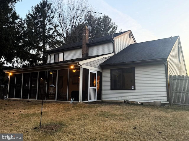 rear view of house with a lawn, a sunroom, a chimney, roof with shingles, and fence