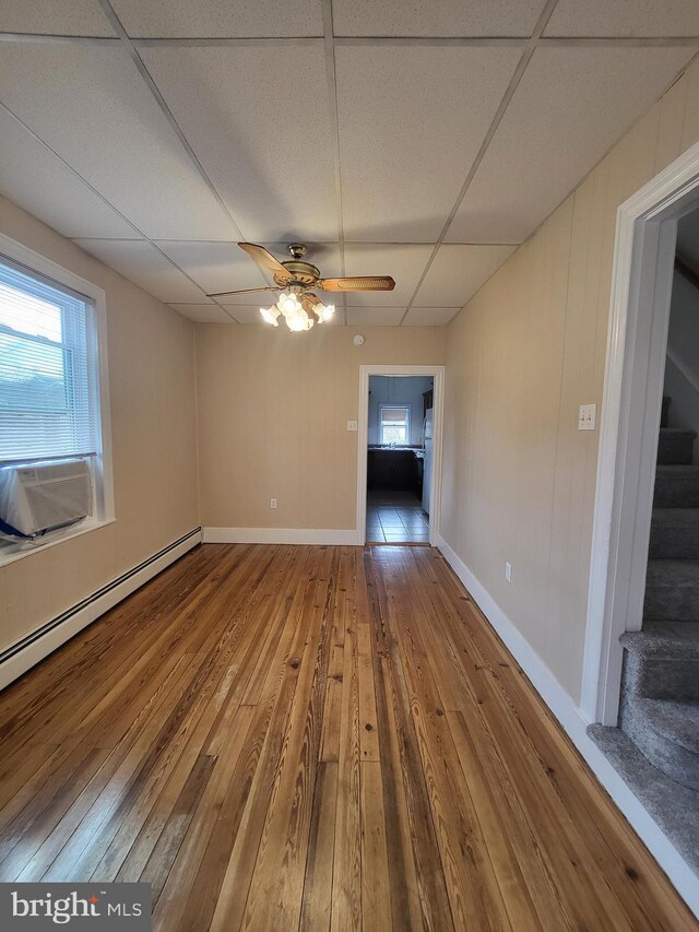 empty room with a ceiling fan, hardwood / wood-style flooring, a baseboard radiator, stairway, and a paneled ceiling