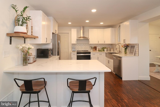 kitchen featuring a peninsula, a breakfast bar, a sink, appliances with stainless steel finishes, and wall chimney exhaust hood