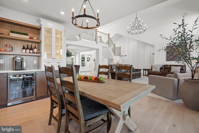 dining room featuring a notable chandelier, beverage cooler, light wood finished floors, a dry bar, and stairs