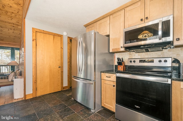 kitchen featuring stainless steel appliances, backsplash, light brown cabinetry, dark countertops, and stone tile flooring