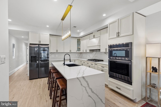 kitchen featuring light wood finished floors, backsplash, custom range hood, stainless steel appliances, and a sink