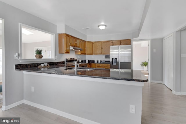 kitchen with under cabinet range hood, a peninsula, appliances with stainless steel finishes, dark stone counters, and brown cabinetry