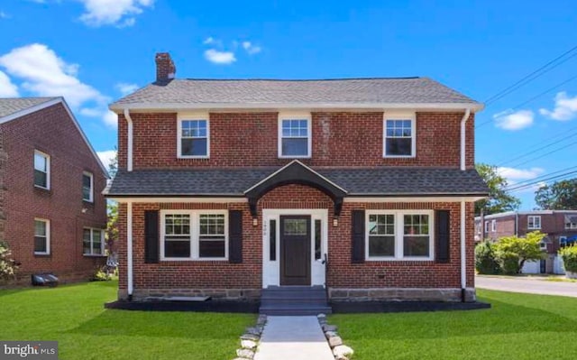 view of front of home with a shingled roof, a front yard, brick siding, and a chimney