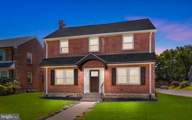 view of front of house with a yard, roof with shingles, a chimney, and brick siding