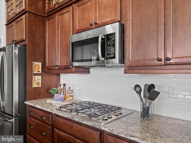 kitchen with appliances with stainless steel finishes, backsplash, light stone countertops, brown cabinetry, and glass insert cabinets