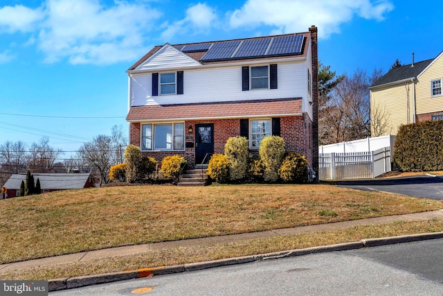 traditional-style home with brick siding, fence, roof mounted solar panels, a front lawn, and a chimney