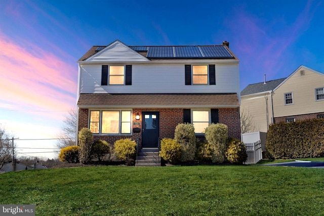 view of front of home featuring a shingled roof, roof mounted solar panels, brick siding, and a yard