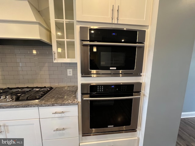 kitchen featuring tasteful backsplash, custom range hood, glass insert cabinets, appliances with stainless steel finishes, and white cabinetry