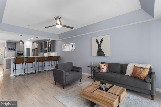 living area with a raised ceiling, a ceiling fan, baseboards, and light wood finished floors