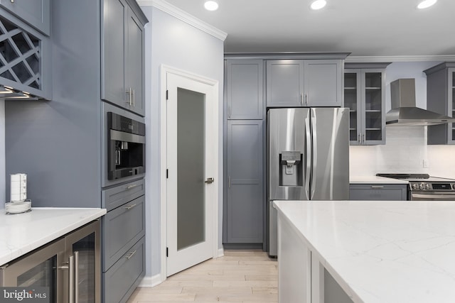 kitchen featuring light stone counters, appliances with stainless steel finishes, wall chimney exhaust hood, crown molding, and glass insert cabinets