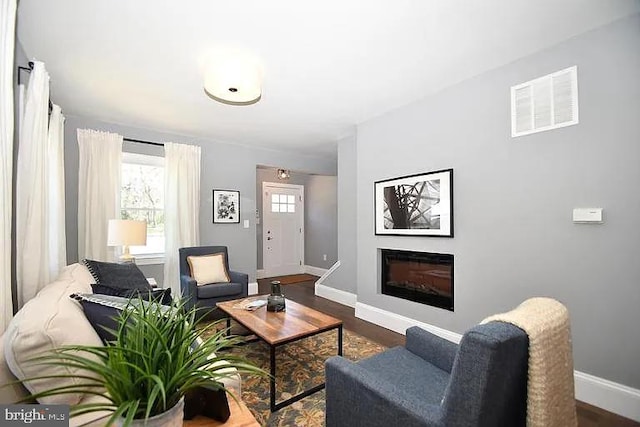 living area with a glass covered fireplace, dark wood-style flooring, visible vents, and baseboards