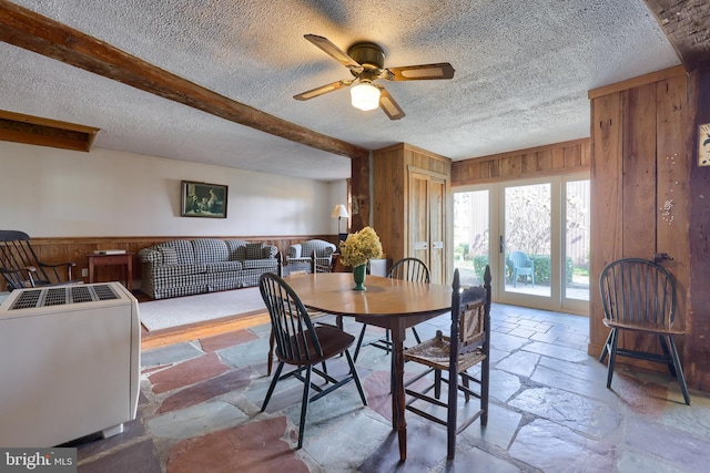 dining space with beam ceiling, stone tile flooring, a ceiling fan, wood walls, and a textured ceiling