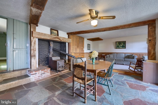 dining area with stone tile floors, wainscoting, beamed ceiling, a wood stove, and a textured ceiling