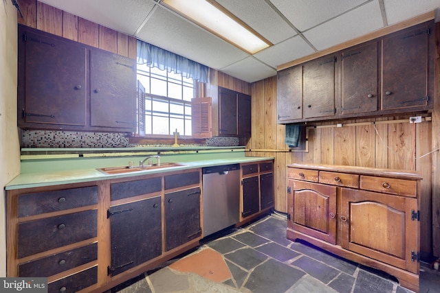 kitchen featuring a drop ceiling, stainless steel dishwasher, light countertops, wood walls, and a sink