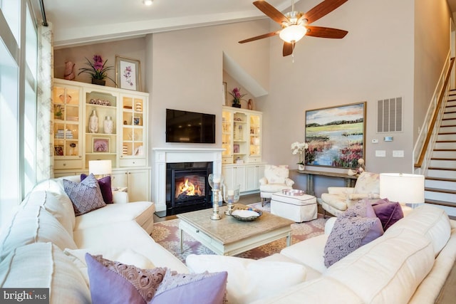 living room with visible vents, a ceiling fan, a glass covered fireplace, stairs, and high vaulted ceiling