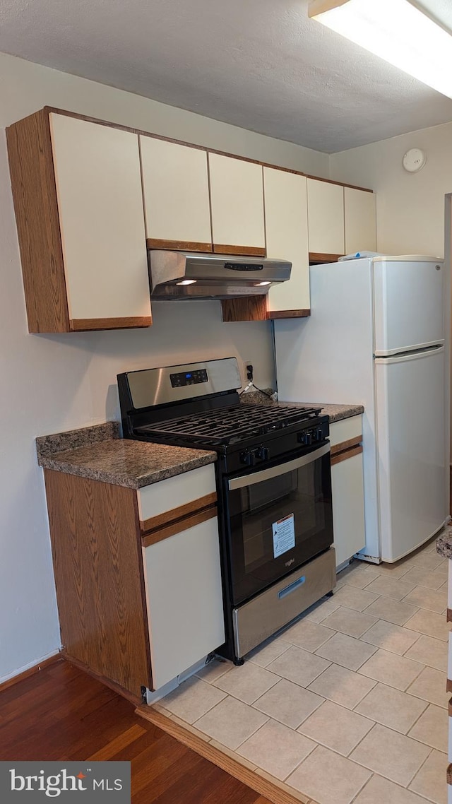 kitchen featuring dark countertops, gas stove, freestanding refrigerator, white cabinetry, and under cabinet range hood