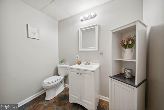 bathroom featuring vanity, baseboards, a drop ceiling, stone finish flooring, and toilet