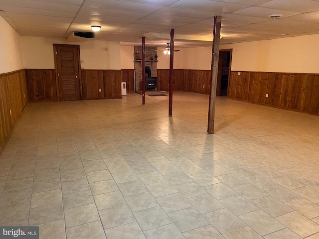 finished basement featuring a paneled ceiling, wood walls, a wood stove, and wainscoting