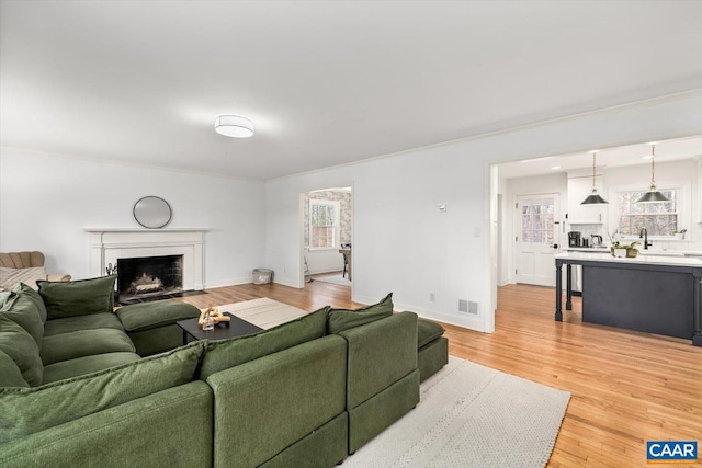 living room with light wood-style flooring, a fireplace with flush hearth, visible vents, baseboards, and ornamental molding