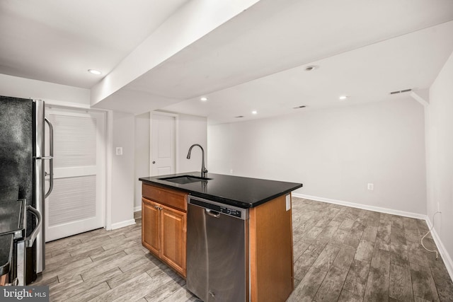 kitchen with stainless steel appliances, dark countertops, light wood-style flooring, a kitchen island with sink, and a sink