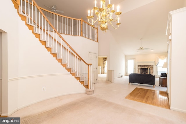 carpeted entryway featuring ceiling fan with notable chandelier, stairway, a fireplace, baseboards, and a towering ceiling