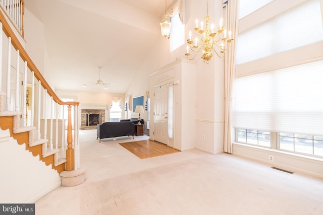 carpeted foyer featuring visible vents, stairway, ceiling fan with notable chandelier, a fireplace, and high vaulted ceiling