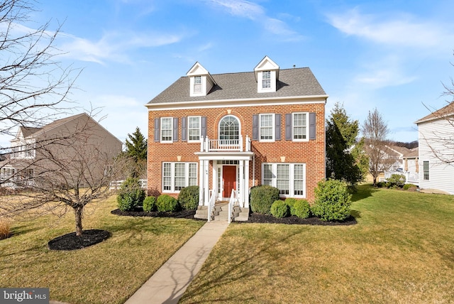 colonial-style house with roof with shingles, brick siding, a front lawn, and a balcony