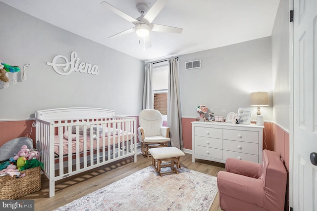 bedroom with a nursery area, visible vents, ceiling fan, and wood finished floors