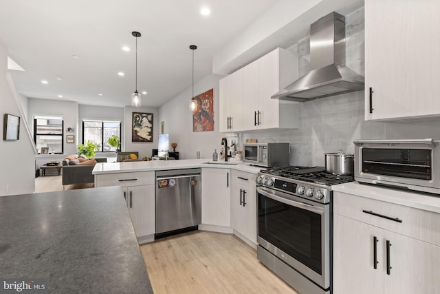 kitchen featuring a peninsula, light wood-style floors, stainless steel appliances, wall chimney exhaust hood, and a sink