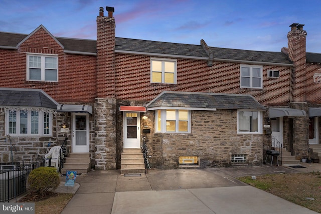 back of house at dusk with entry steps, stone siding, and a chimney