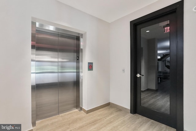 corridor featuring elevator, light wood-style flooring, and baseboards